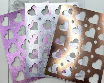 May include: Three sheets of decorative paper in shades of purple, silver, and copper. Each sheet features a pattern of heart-shaped cutouts and small circular perforations. The paper has a metallic sheen, creating a shimmering effect.