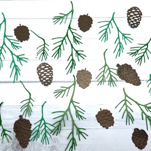 May include: Green paper pine branches and brown paper pine cones on a white wooden background.