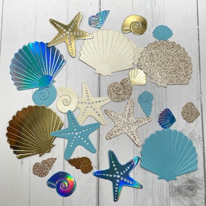 May include: A collection of decorative seashells in various colors and textures, including gold, blue, white, and brown. The shells are arranged on a white wooden surface.