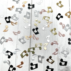 May include: A scattering of silver, gold, and black musical note confetti on a white wooden surface. The notes are in various sizes and shapes.