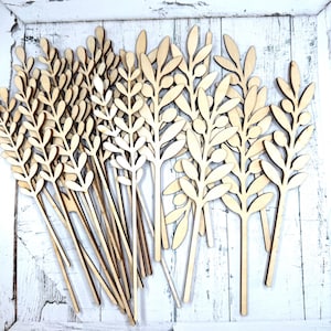 May include: A collection of light-colored wooden cutouts shaped like olive branches with leaves and small round fruits. Each piece features a long, thin stem attached to a detailed botanical silhouette, crafted from flat, smooth, natural wood.