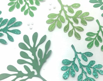 May include: An assortment of green leaf-shaped embellishments in various shades, including glittery teal and matte olive. Small, pearl-like beads are scattered among the leaves. The leaves are arranged on a white surface, with a ruler visible.