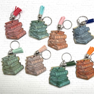 May include: A collection of keychains featuring stacks of books in various colors, including red, blue, green, and orange. Each keychain has a matching tassel. The keychains are on a white brick background.