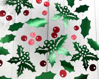 May include: Green holly leaves and red berry confetti scattered on a white surface. The metallic green leaves have detailed veins, and the red berries are small and round. Festive holiday decorations.
