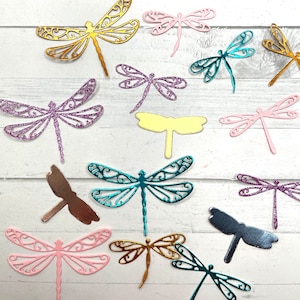 May include: A collection of colorful dragonfly cutouts in various shades, including gold, pink, teal, purple, and brown. The dragonflies are arranged on a white wooden surface, showcasing their intricate wing designs and delicate forms. These decorative elements are ideal for crafting and scrapbooking.