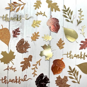 May include: An assortment of fall-themed die-cuts in various metallic finishes. Includes leaves, acorns, squirrels, and the word "thankful" in gold. Some elements have glitter accents, creating a festive autumn aesthetic.