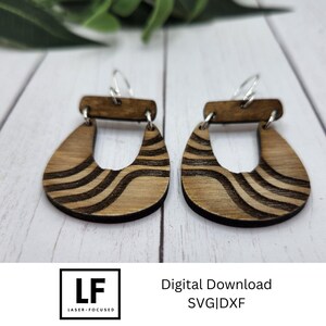 May include: A pair of wooden earrings with a wavy pattern. The earrings are brown with black lines and have a simple hoop design. The earrings are shown on a white surface.