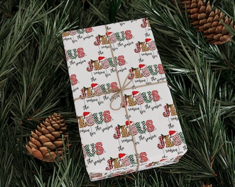 May include: A wrapped gift with a white background featuring the text "JESUS is the reason for the season" in red, green, and leopard print. The gift is tied with twine and rests on a bed of pine needles and pine cones.