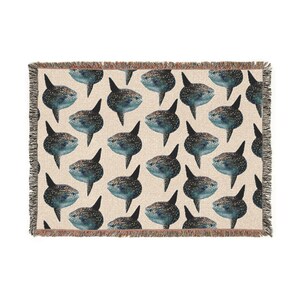 May include: A woven throw blanket featuring a repeating pattern of dark blue and grey sunfish on a light beige background. The blanket has a textured surface and finished edges with a short, multi-colored fringe along all four sides.