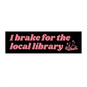 Puede incluir: Una pegatina rectangular negra con el texto rosa "I brake for the local library". Presenta una ilustración de un libro abierto y estrellas. El fondo es blanco.