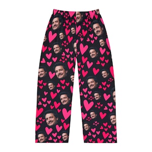 May include: Black lounge pants featuring an all-over repeating pattern of bright pink hearts and a repeated photographic portrait of a man with dark hair and a beard. The pants have a straight-leg cut and an elasticized waistband at the top.