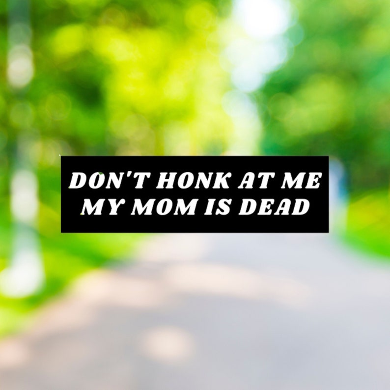May include: A black and white sign with the text "DON'T HONK AT ME MY MOM IS DEAD" against a blurred green background.