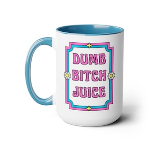 May include: A white ceramic mug with a blue handle and interior rim. The front features a rectangular frame in pink and blue with yellow star accents. Inside the frame, the text DUMB BITCH JUICE is printed in a bold, pink, wavy font.