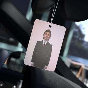 May include: A rectangular car air freshener featuring a printed image of a person with dark hair wearing a black suit, a white collared shirt, and a striped tie. The air freshener is suspended by a thin white elastic string from a rearview mirror.
