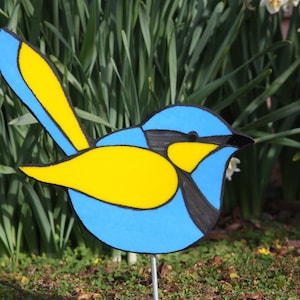 Peut inclure: Décoration d'oiseau en bois peint, avec un corps bleu, des ailes jaunes et des détails noirs. L'oiseau est monté sur une tige métallique, idéal pour un jardin. Le fond présente un feuillage vert.