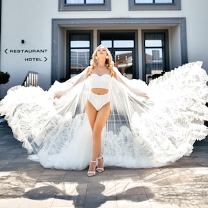 Op de afbeelding: Een vrouw draagt een witte bruidslingerie set met een lange, vloeiende tule cape. De cape heeft een gerimpeld ontwerp en is achter haar uitgespreid. Ze staat voor een gebouw met het bord "Restaurant Hotel" erop.