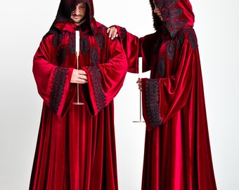 May include: Two people wearing long, red velvet robes with black lace trim and hoods. They are holding candles in their hands.