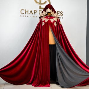 May include: A deep red velvet hooded cloak with gold lace trim and a gray lining. The cloak is displayed on a mannequin, with the words "CHAP DRESSES CREATING STYLE" visible in gold lettering on the wall behind it.