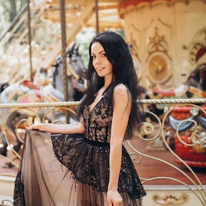 May include: A floor-length dress featuring a black lace bodice with a deep V-neckline over a nude-colored lining. The skirt consists of a layered black tulle overlay with sequin embellishments and a solid black waistband, creating a full silhouette.