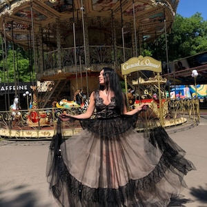 May include: A floor-length dress featuring a black lace bodice with a V-neckline and a voluminous skirt. The skirt consists of a beige tulle base layered with black tulle ruffles and intricate black lace trim along the hem and waistline.