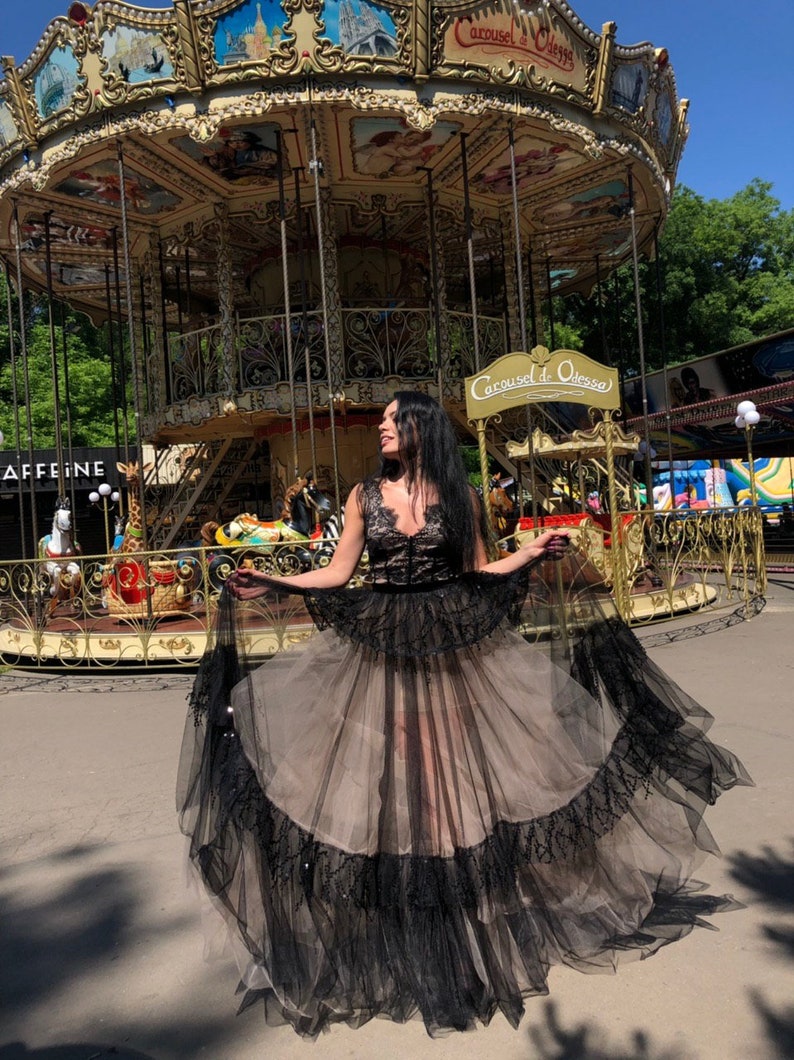May include: A floor-length dress featuring a black lace bodice with a V-neckline and a voluminous skirt. The skirt consists of a beige tulle base layered with black tulle ruffles and intricate black lace trim along the hem and waistline.