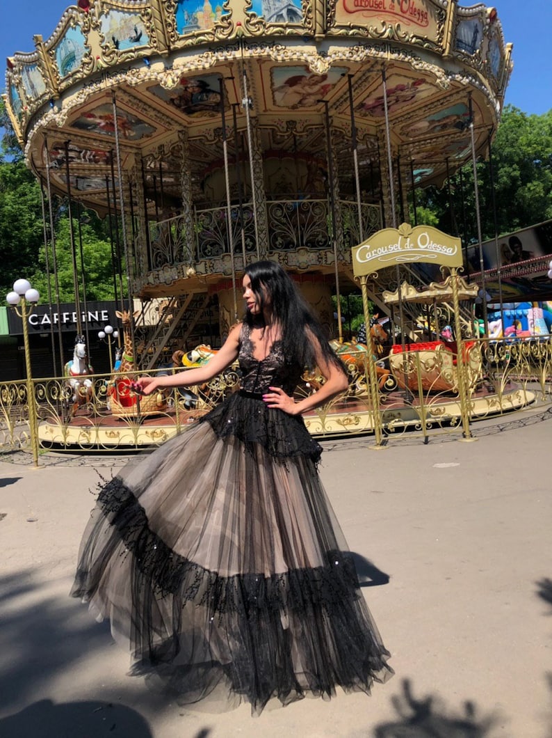 May include: A floor-length gown featuring a black lace bodice with a V-neckline and a layered skirt. The skirt consists of a beige tulle base with black tulle overlays, accented by black lace trim and scattered sequins throughout the fabric.