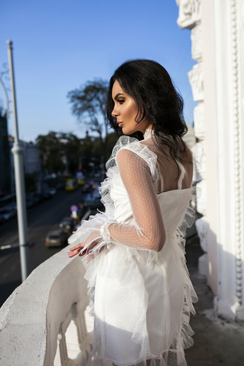 May include: A woman wearing a white lace dress with ruffles and a sheer overlay. She is standing on a balcony overlooking a city street.