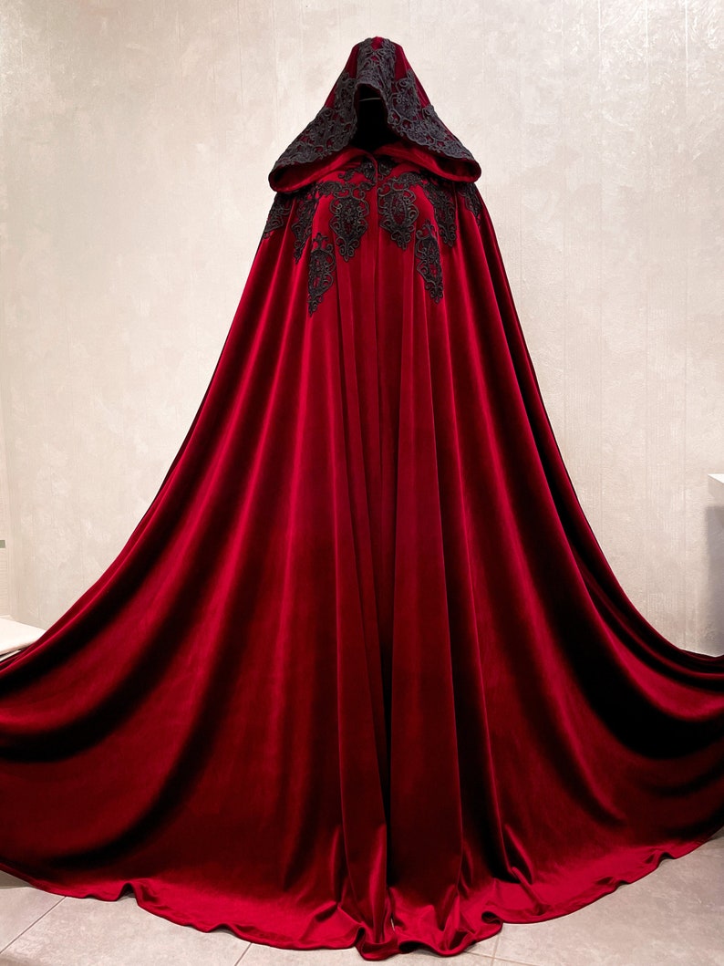 May include: A long, flowing red velvet cape with a black lace trim and hood. The cape is draped over a mannequin and has a dramatic, elegant look.