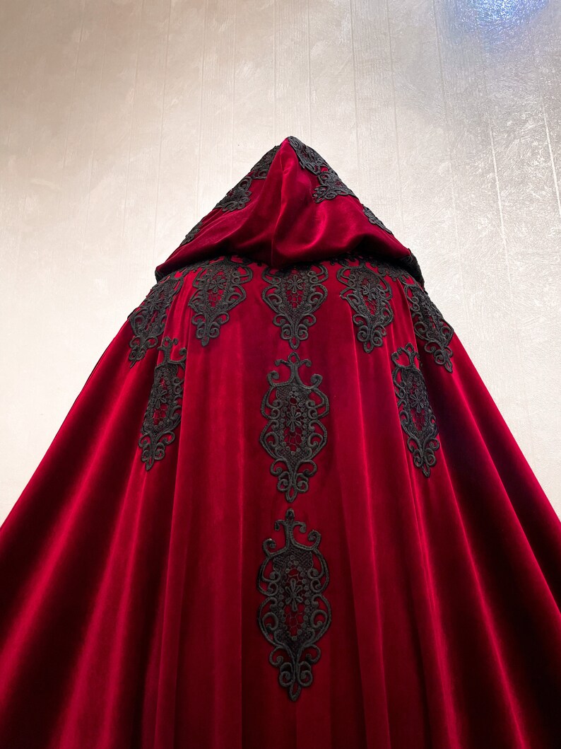 May include: A red velvet hooded cape with black lace trim. The cape has a flowing design and is perfect for a dramatic look.