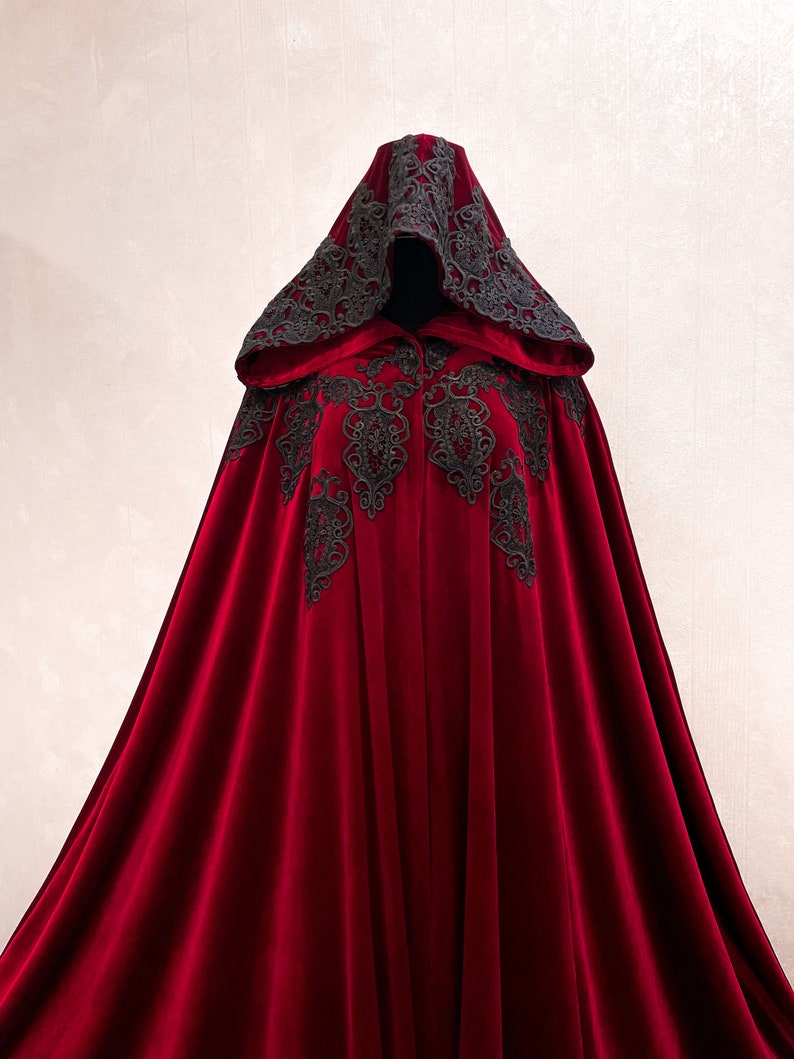 May include: A long, red velvet cape with a black lace trim and hood. The lace trim features intricate floral designs.