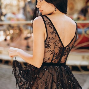 May include: A black sleeveless dress featuring a sheer lace bodice with floral patterns and a V-neckline. The waist is defined by a solid black band, and the skirt is adorned with intricate black sequin embroidery and dangling sequin accents.