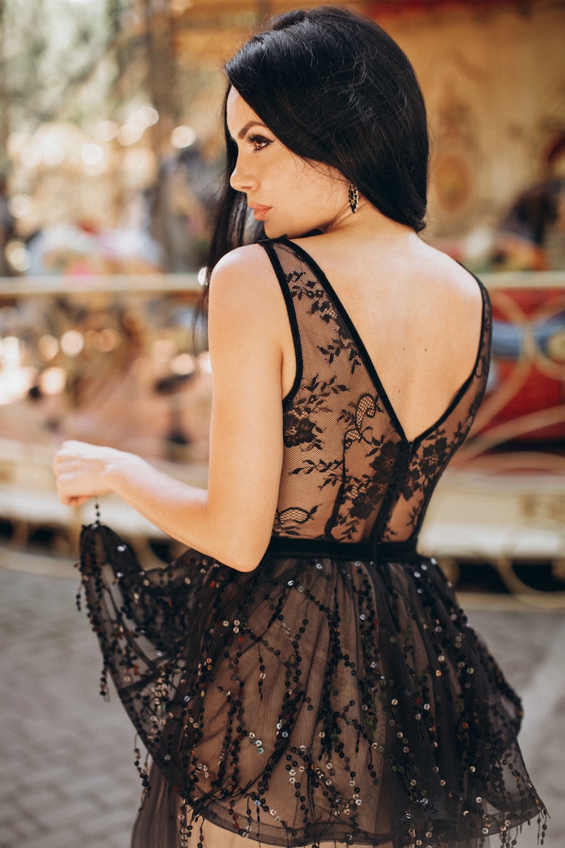May include: A black sleeveless dress featuring a sheer lace bodice with floral patterns and a V-neckline. The waist is defined by a solid black band, and the skirt is adorned with intricate black sequin embroidery and dangling sequin accents.