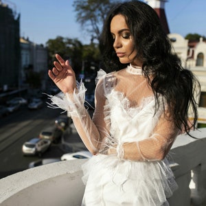 May include: A woman wearing a white lace dress with a sheer overlay and ruffled details. She is standing on a balcony overlooking a city street.