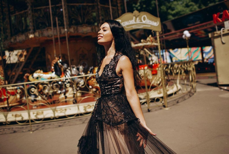 May include: A sleeveless black dress featuring a lace bodice with a V-neckline and a layered tulle skirt. The bodice is adorned with intricate black embroidery and sequins, while the skirt consists of sheer black tulle over a neutral lining.