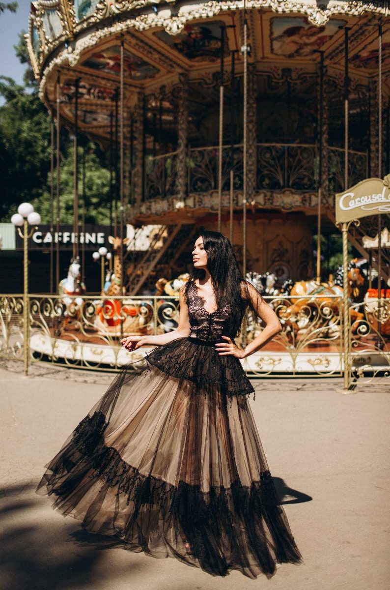 May include: A floor-length gown featuring a black lace bodice with a deep V-neckline and a layered skirt. The skirt consists of a beige underskirt covered by a sheer black tulle overlay with a wide black lace trim along the bottom hem.