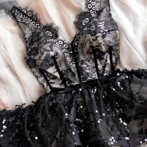 May include: A sleeveless bodice featuring black floral lace over a beige lining with a deep V-neckline and vertical velvet trim. The waist is accented by a black velvet band, leading to a flared peplum skirt covered in black sequins.