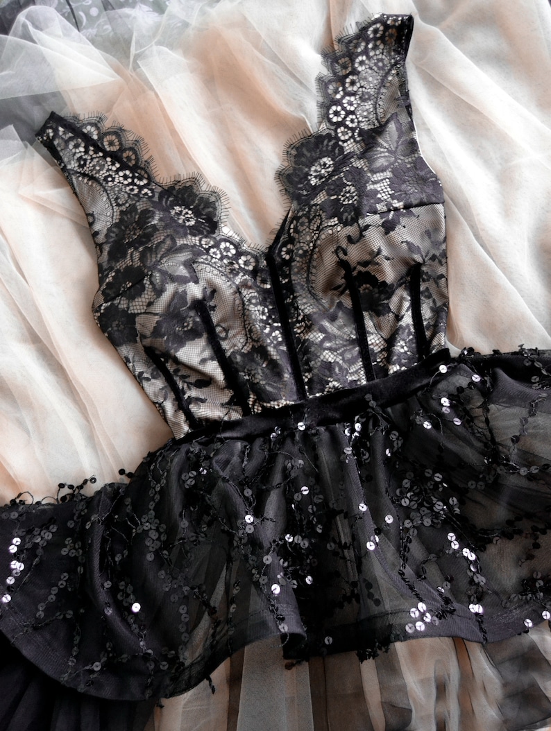 May include: A sleeveless bodice featuring black floral lace over a beige lining with a deep V-neckline and vertical velvet trim. The waist is accented by a black velvet band, leading to a flared peplum skirt covered in black sequins.