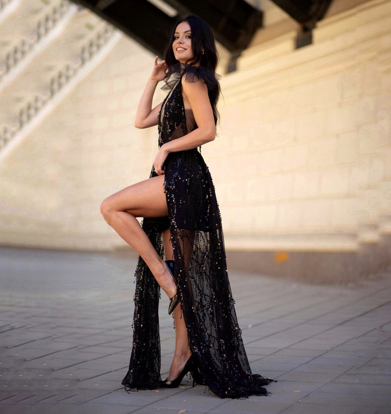 Puede incluir: Una mujer con un vestido negro de lentejuelas con una superposici&oacute;n transparente y una abertura alta. Est&aacute; de pie frente a una pared blanca y lleva tacones altos negros.