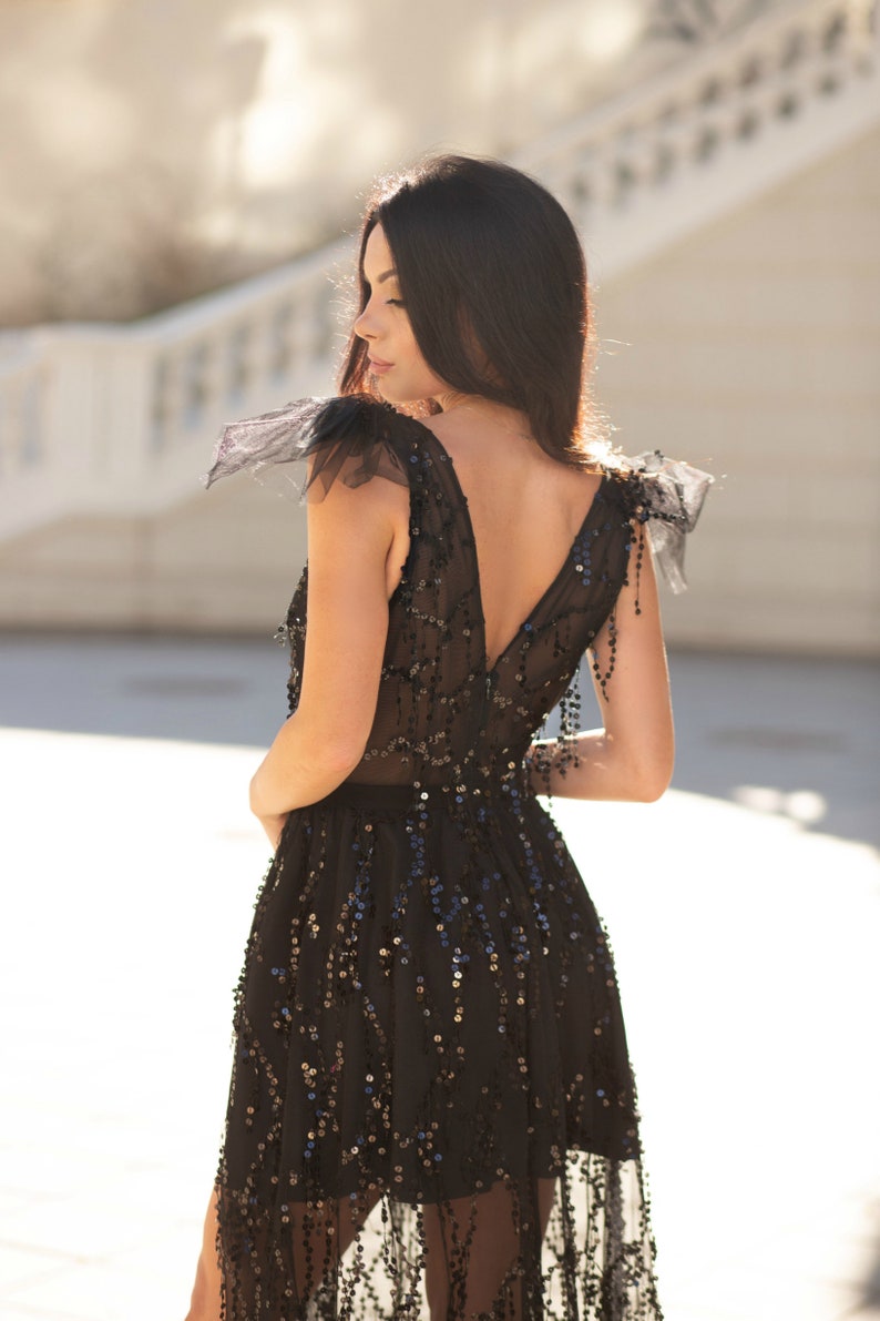 Puede incluir: Una mujer con un vestido negro de lentejuelas con mangas transparentes y un escote pronunciado. El vestido tiene una falda fluida y est&aacute; adornado con lentejuelas.
