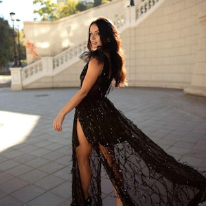 Puede incluir: Una mujer con un vestido negro de lentejuelas con una superposici&oacute;n transparente y una abertura alta. El vestido tiene una cola fluida y la mujer est&aacute; de pie sobre una superficie pavimentada.