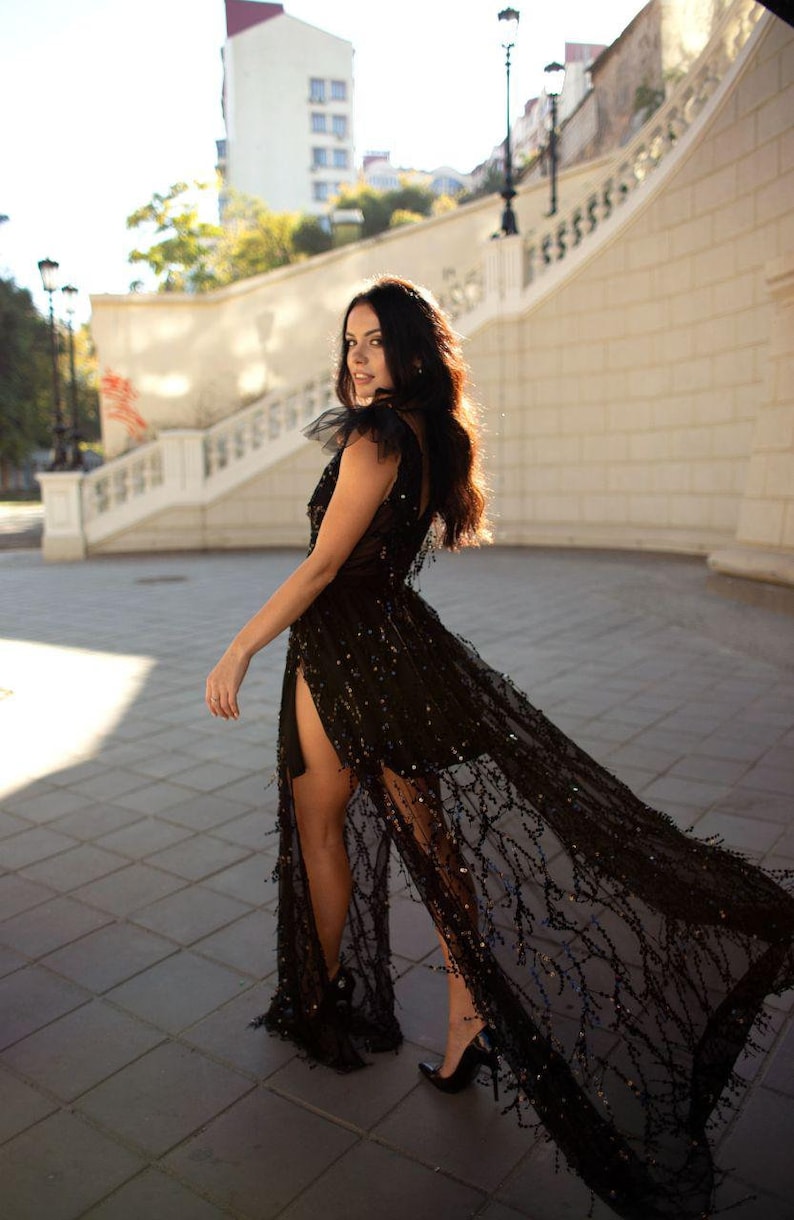 Puede incluir: Una mujer con un vestido negro de lentejuelas con una superposici&oacute;n transparente y una abertura alta. El vestido tiene una cola fluida y la mujer est&aacute; de pie sobre una superficie pavimentada.