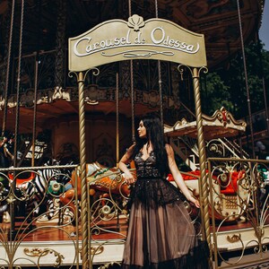 May include: A floor-length black dress with a lace bodice and a layered tulle skirt, featuring a sheer overlay and intricate embroidery. The garment is positioned in front of a gold-toned carousel sign that reads Carousel de Odessa.