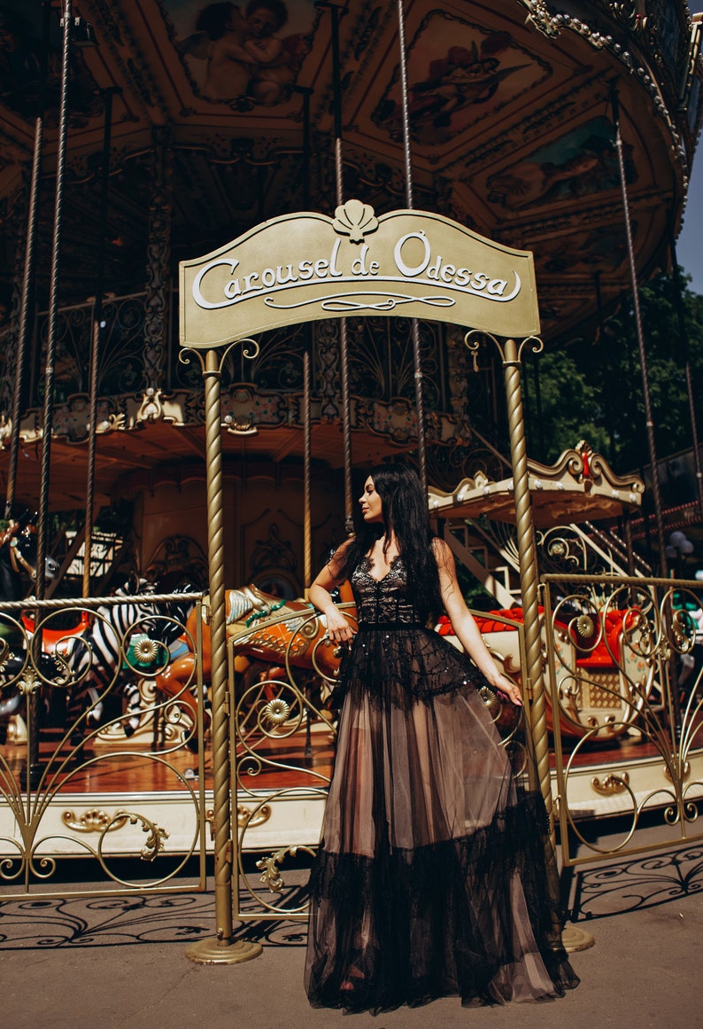 May include: A floor-length black dress with a lace bodice and a layered tulle skirt, featuring a sheer overlay and intricate embroidery. The garment is positioned in front of a gold-toned carousel sign that reads Carousel de Odessa.