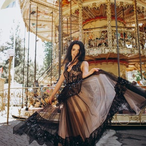 May include: A sleeveless floor-length dress featuring a nude-colored base layer with a black lace overlay. The bodice has a V-neckline and intricate black lace detailing, while the skirt consists of a sheer black tulle overlay with a wide, decorative black lace hemline.