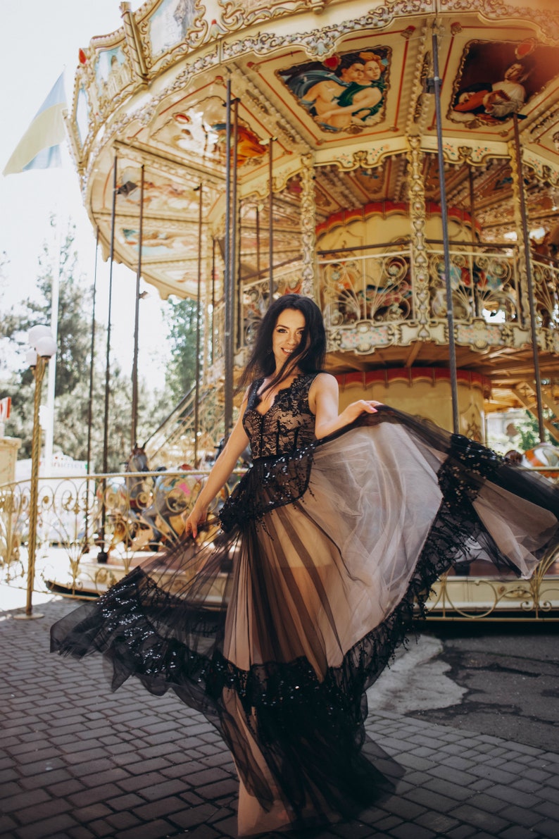 May include: A sleeveless floor-length dress featuring a nude-colored base layer with a black lace overlay. The bodice has a V-neckline and intricate black lace detailing, while the skirt consists of a sheer black tulle overlay with a wide, decorative black lace hemline.