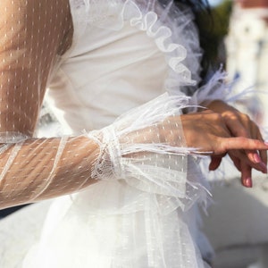 May include: A white dress with a sheer, dotted fabric and ruffled detailing. The dress has long sleeves with feather trim and a fitted bodice.