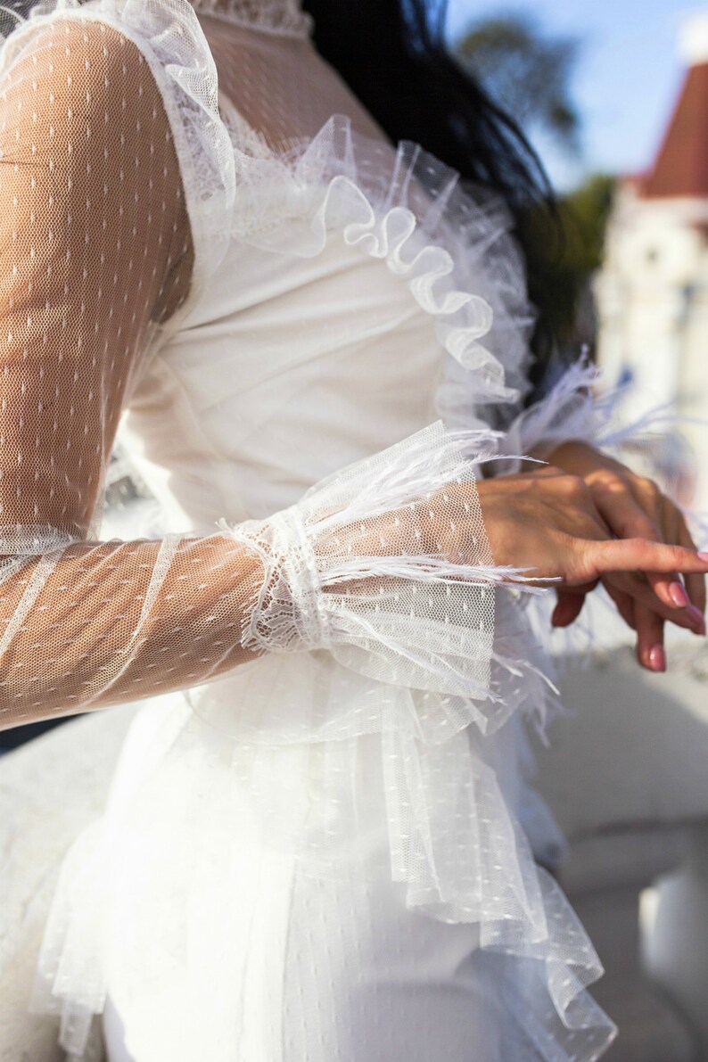 May include: A white dress with a sheer, dotted fabric and ruffled detailing. The dress has long sleeves with feather trim and a fitted bodice.