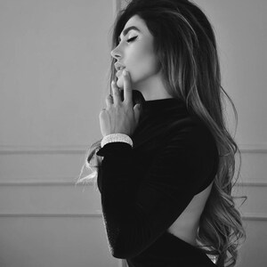 K&ouml;nnte beinhalten: Eine Frau mit langen, dunklen Haaren tr&auml;gt ein schwarzes Samtkleid mit tiefem R&uuml;cken. Sie schaut nach unten und ber&uuml;hrt ihr Kinn mit der Hand. Sie tr&auml;gt ein silbernes Armband am Handgelenk.