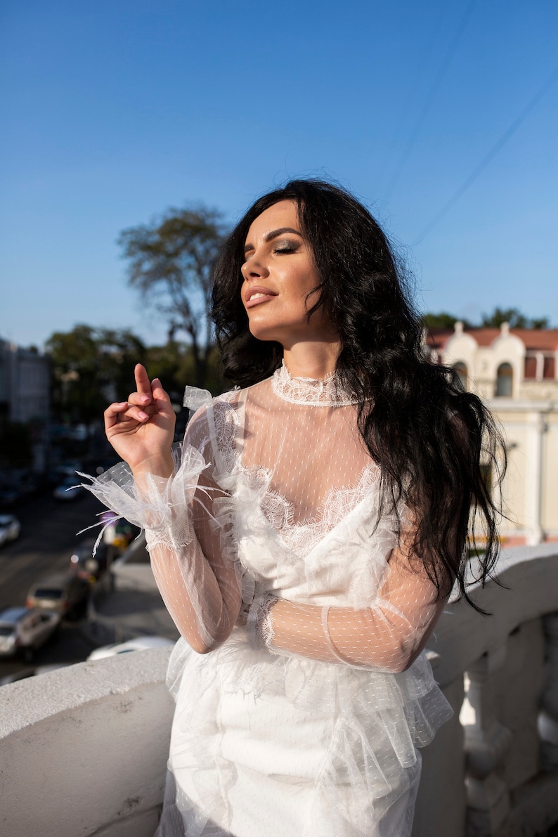 May include: A woman with long black hair wearing a white lace dress with a sheer overlay. She is standing on a balcony with a city view in the background.