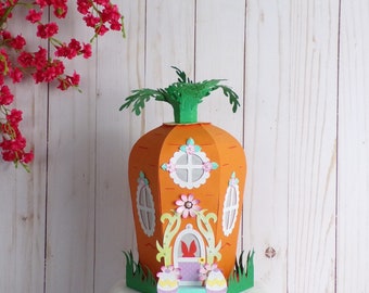 May include: A papercraft carrot house with a green leafy top, a white door with a pink bunny, and two yellow and blue Easter eggs. The house is decorated with flowers and windows.